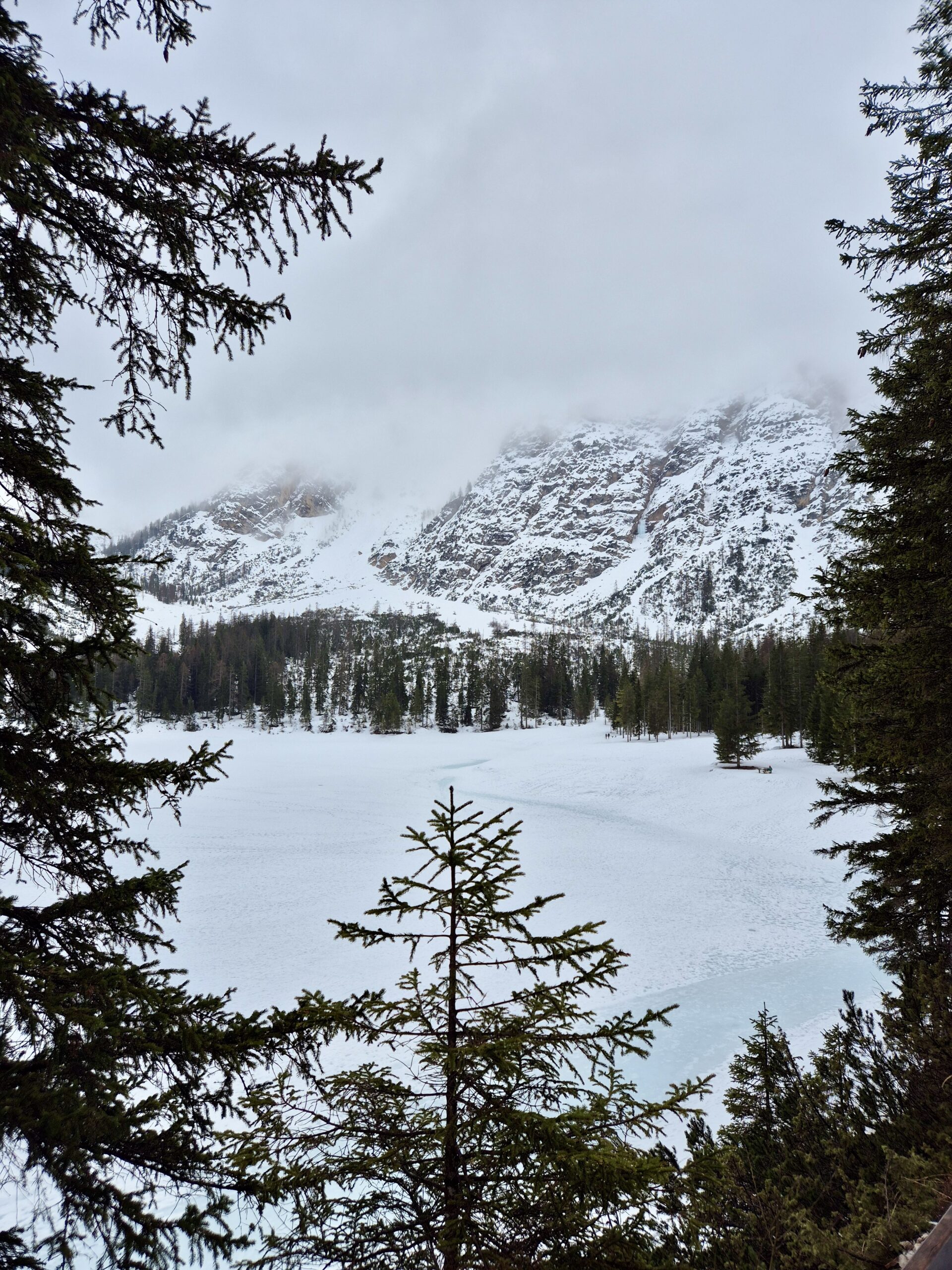 Verschneite Berglandschaft mit gefrorenem See Ein winterliches Naturpanorama mit einem zugefrorenen See, umgeben von schneebedeckten Bergen und dichten Nadelwäldern. Nebel verhüllt teilweise die Gipfel.