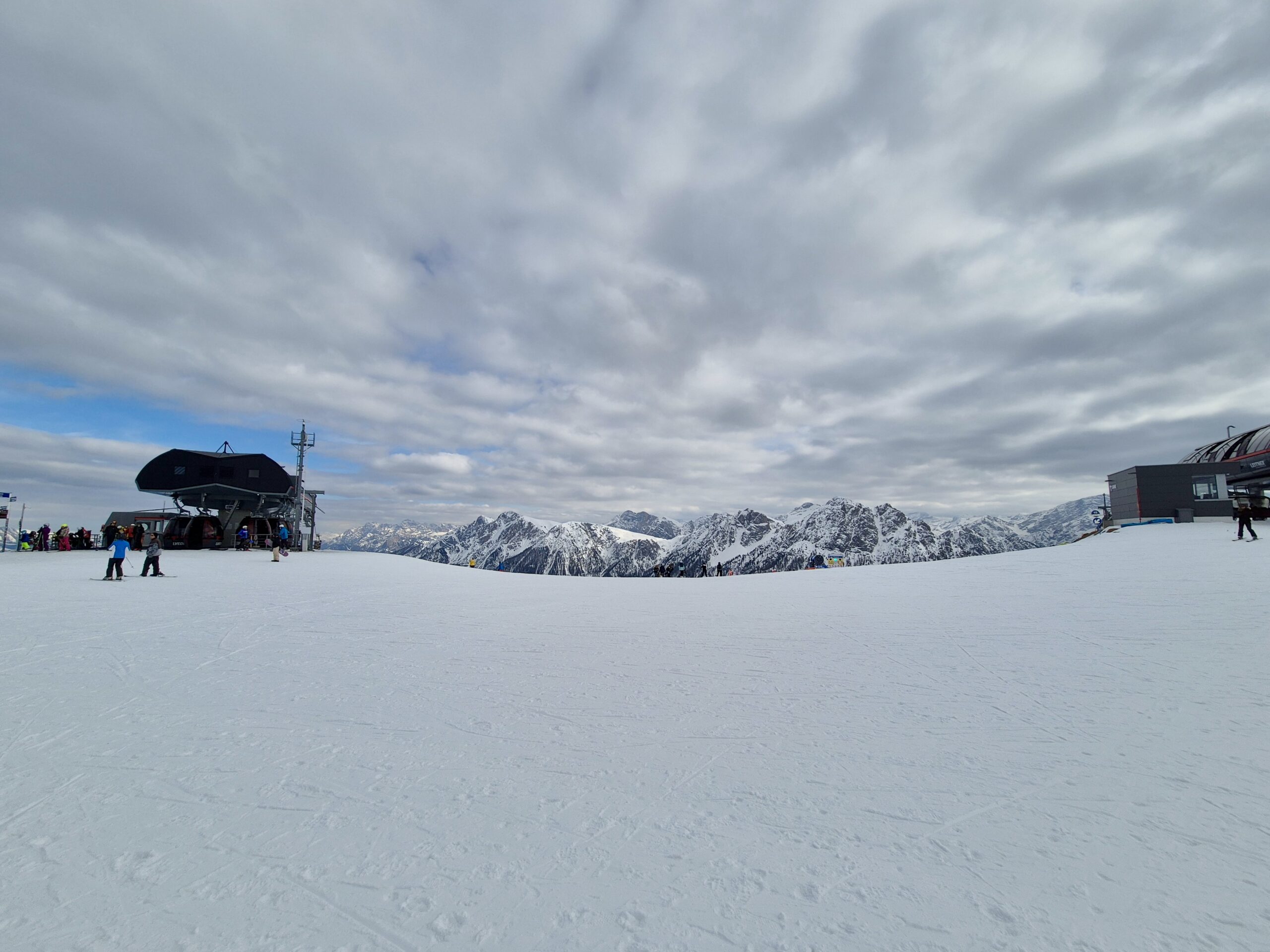 Kronplatz Eine winterliche Berglandschaft mit einer breiten, schneebedeckten Skipiste. Im Hintergrund sind verschneite Gipfel unter einer bewölkten Himmel zu sehen. Zwei moderne Sesselliftstationen befinden sich links und rechts, während Skifahrer und Snowboarder die Piste betreten.